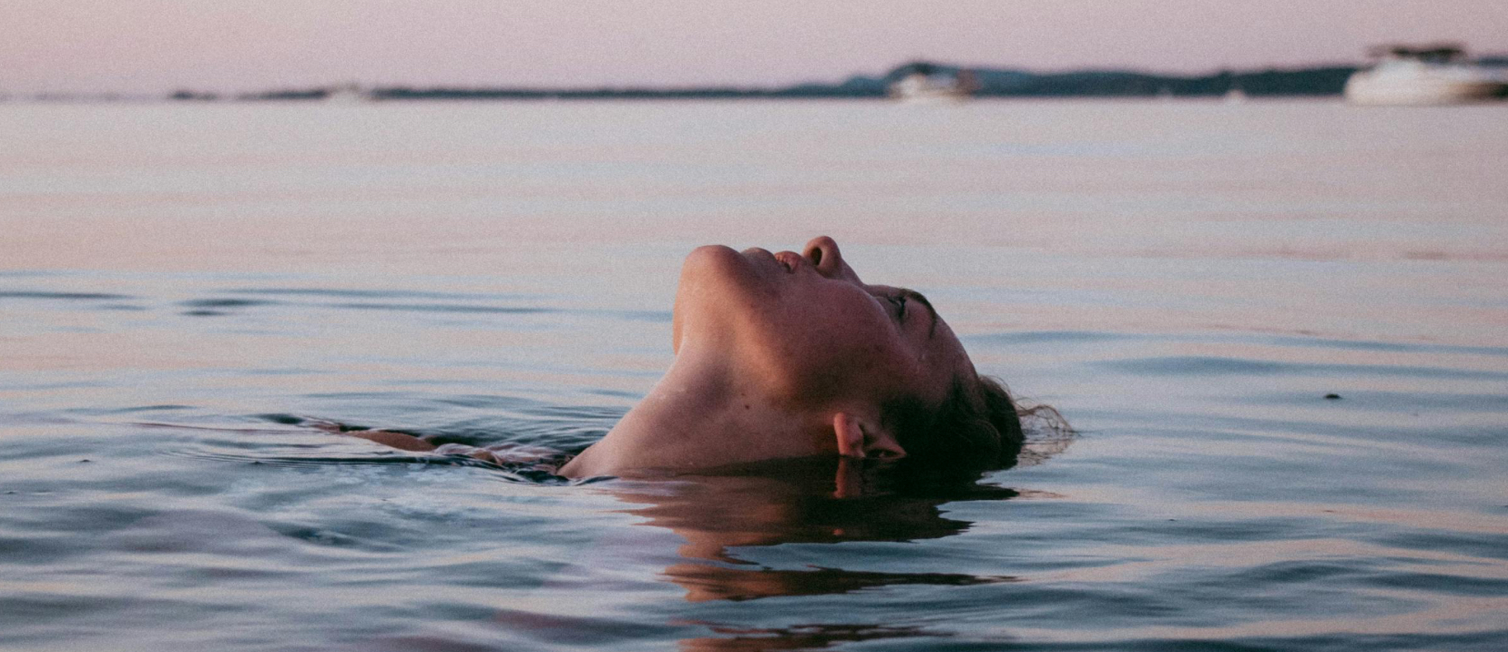 A woman floating on the surface of calm water at dusk — softness and surrender.