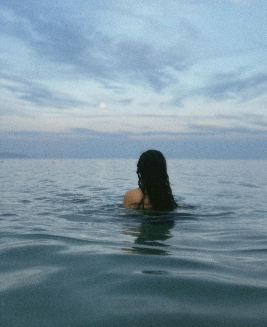 A woman in the ocean at dusk beneath the moon.